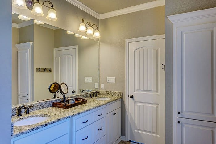 A modern bathroom featuring a double vanity with granite countertops, white cabinetry, a mirror, and a door leading to another room.