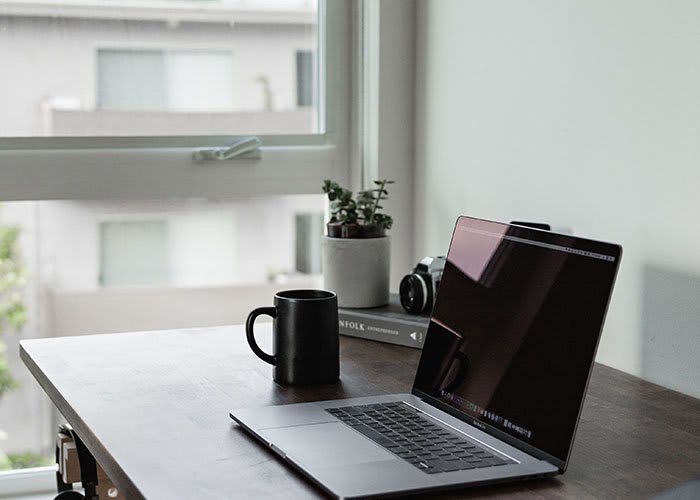 A minimalist workspace featuring a laptop, a coffee mug, and a small plant, with a view of a window and buildings outside.